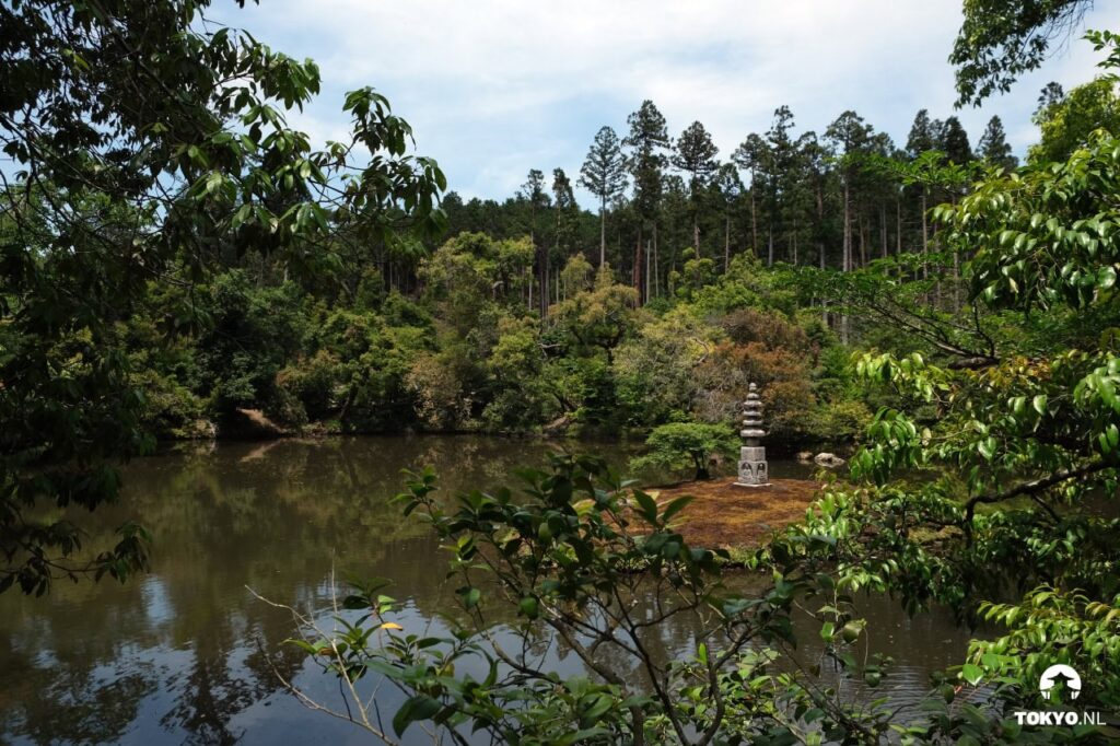 A serene view across a pond showing forested hills in the background, with a traditional stone pagoda or monument visible among the trees on a small island or peninsula.