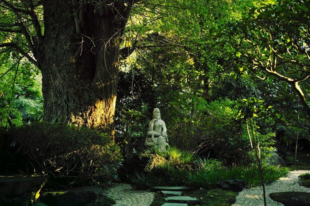 Friedliche Buddha-Statue in einem üppig grünen japanischen Waldgarten mit alten Bäumen und natürlichen Steinwegen
