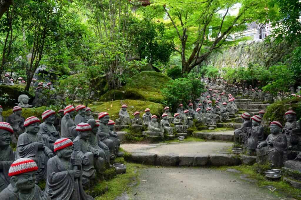 Hunderte kleine Jizo-Buddha-Statuen mit roten Häkelmützen im grünen Wald eines japanischen Tempels bei Hiroshima