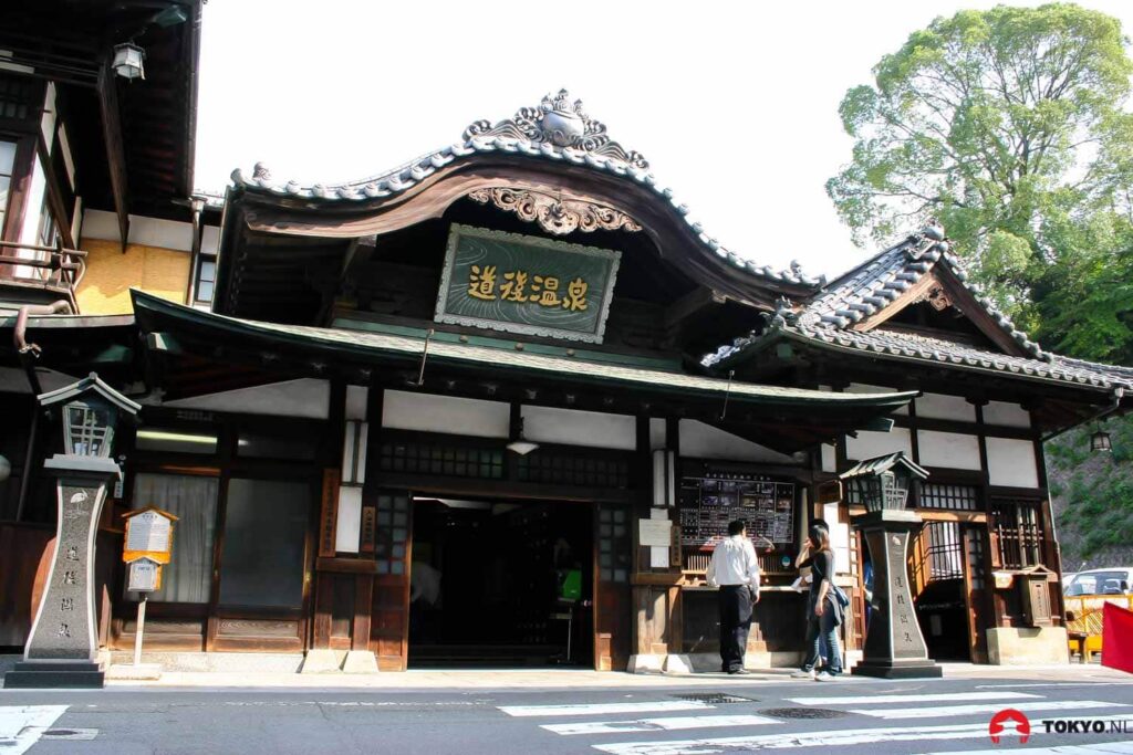 Traditionelles japanisches Onsen-Badehaus Dogo mit geschwungenen Dachziegeln, Holzarchitektur und Besuchern am Eingang in Matsuyama