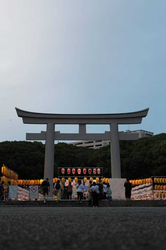 Großes weißes Torii-Tor mit Laternen bei Dämmerung und Festivalbesuchern