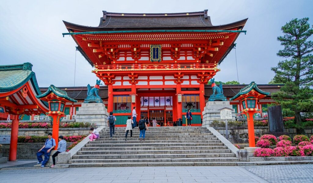 Außenfassade vom Fushimi-Inari Taisha mit roten Akzenten und Steintreppe und Personen. 