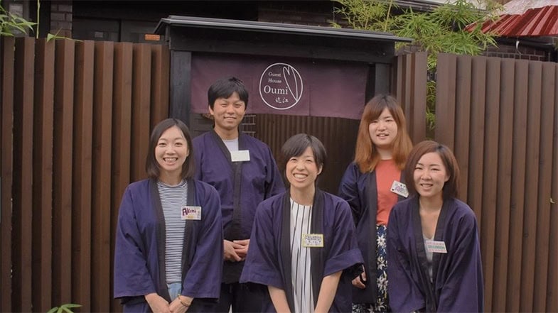 Fünf lächelnde japanische Hotelmitarbeiter in traditionellen dunkelblauen Kimonos stehen vor dem Guest House Oumi Eingang