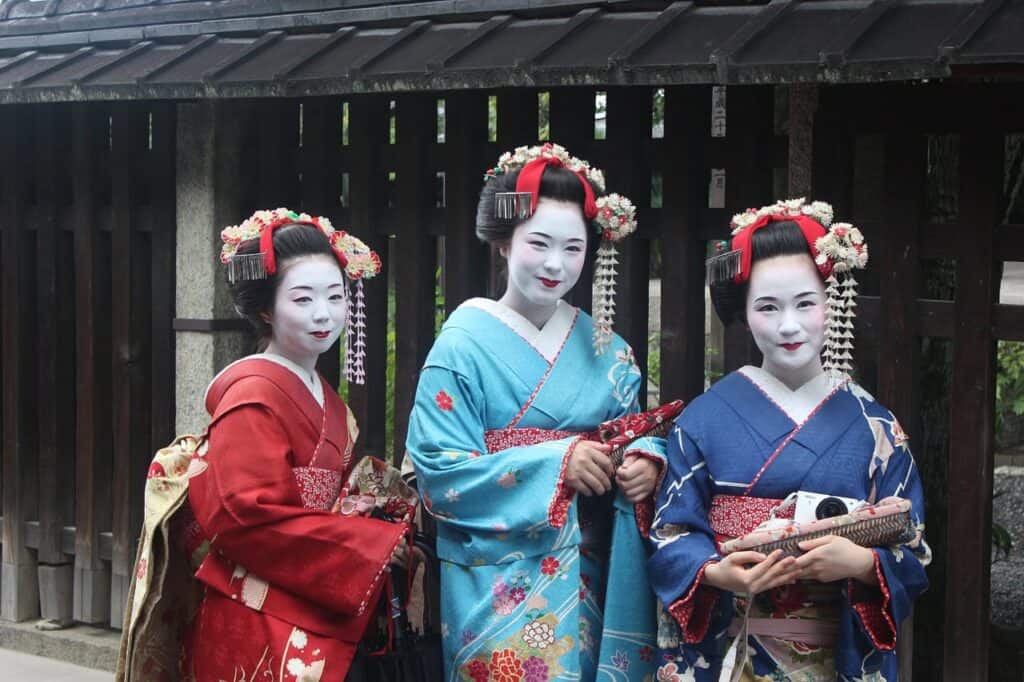 Drei Geishas in traditioneller Kleidung und Make-up. Sie tragen farbenfrohe Kimonos in Rot, Türkis und Blau mit aufwendigen Haarschmuck und den charakteristischen weißen Gesichtern der Geisha-Tradition