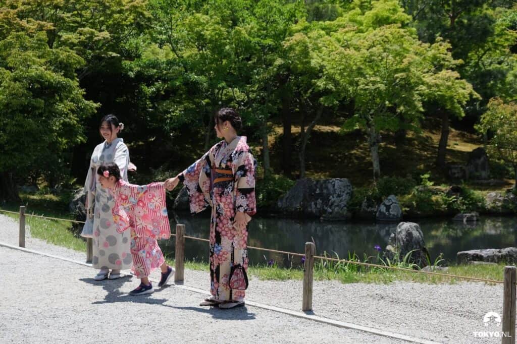 Eine Familie in Kimonos in einem japanischen Garten. Eine Mutter mit ihrem Kind und einer anderen Frau, alle in traditioneller Kleidung mit verschiedenen Mustern und Farben.