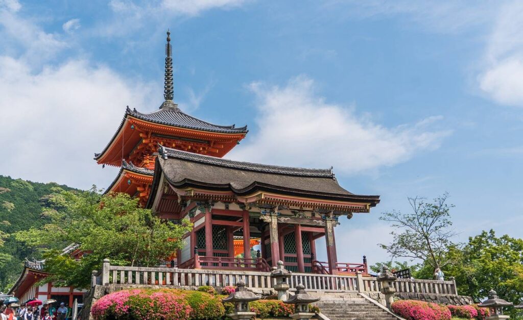 Detailansicht der charakteristischen rot-weißen Pagoda des Kiyomizu-dera mit traditioneller japanischer Dacharchitektur vor blauem Himmel und Bergkulisse