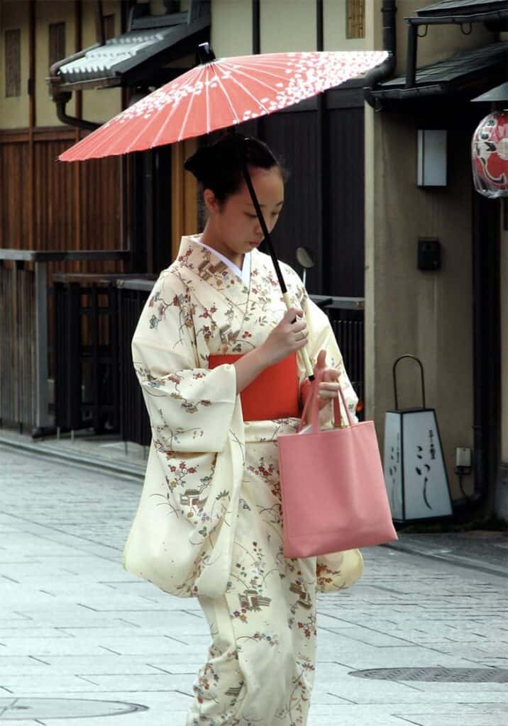 Eine moderne Frau in einem hellen Kimono mit zartem Blumenmuster, die mit einem traditionellen japanischen Sonnenschirm durch eine alte Straße spaziert.
