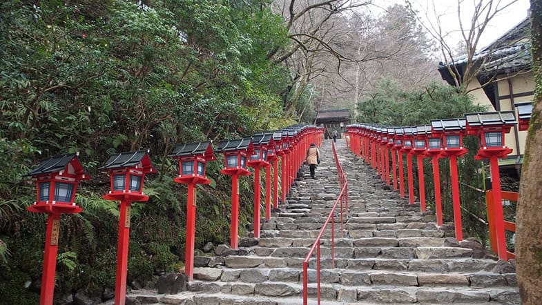 Steintreppe mit roten Laternen führt bergauf zu einem japanischen Schrein, gesäumt von traditionellen Tōrō-Laternen
