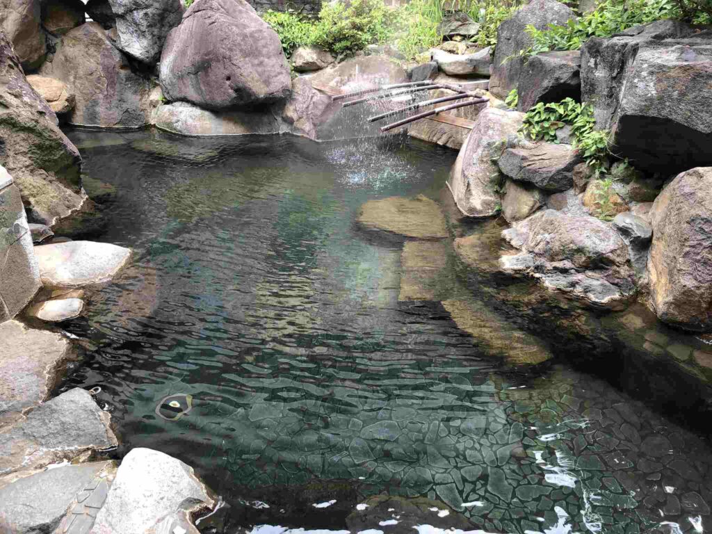 Naturbelassenes Outdoor-Onsen mit kristallklarem Quellwasser, vulkanischen Gesteinen und traditioneller Bambusschöpfkelle in japanischer Berglandschaft