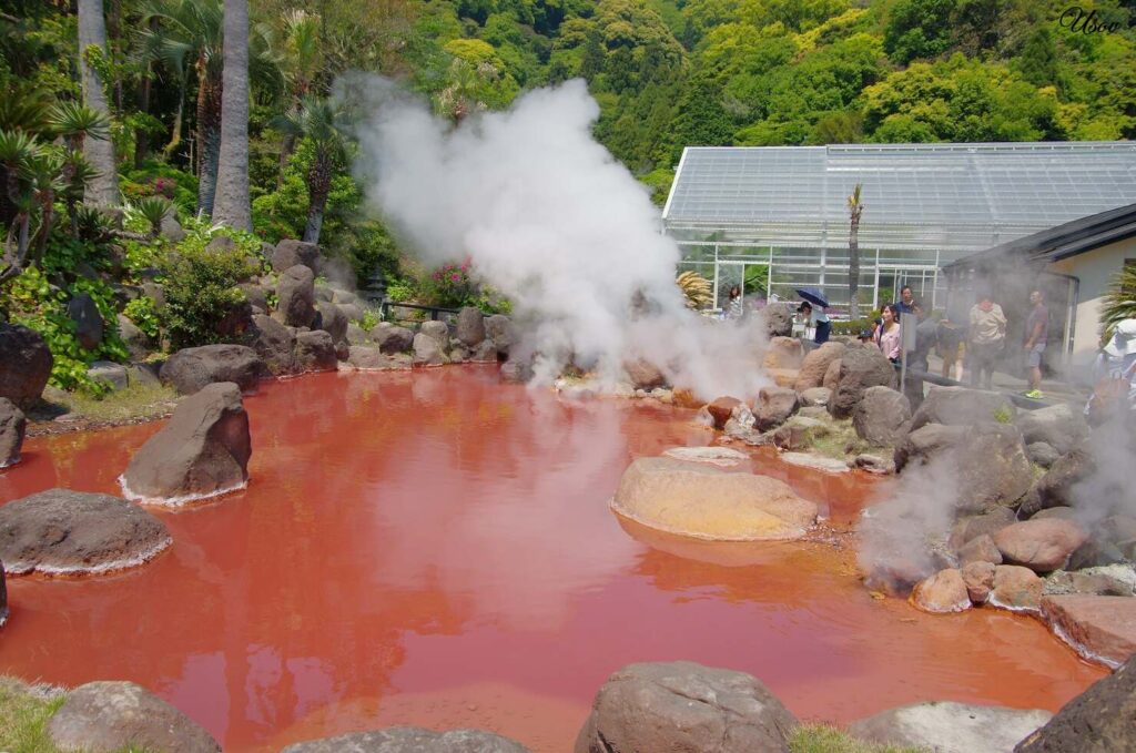 Spektakuläres rotes Onsen-Becken mit eisenhaltigen Mineralien, dampfenden heißen Quellen und Besuchern in tropischer Gartenanlage