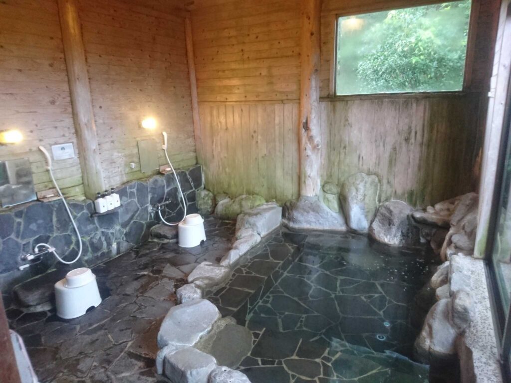 Modernes japanisches Onsen-Innenbad mit dunklen Natursteinfliesen, Holzverkleidung und großem Fenster zur grünen Gartenlandschaft