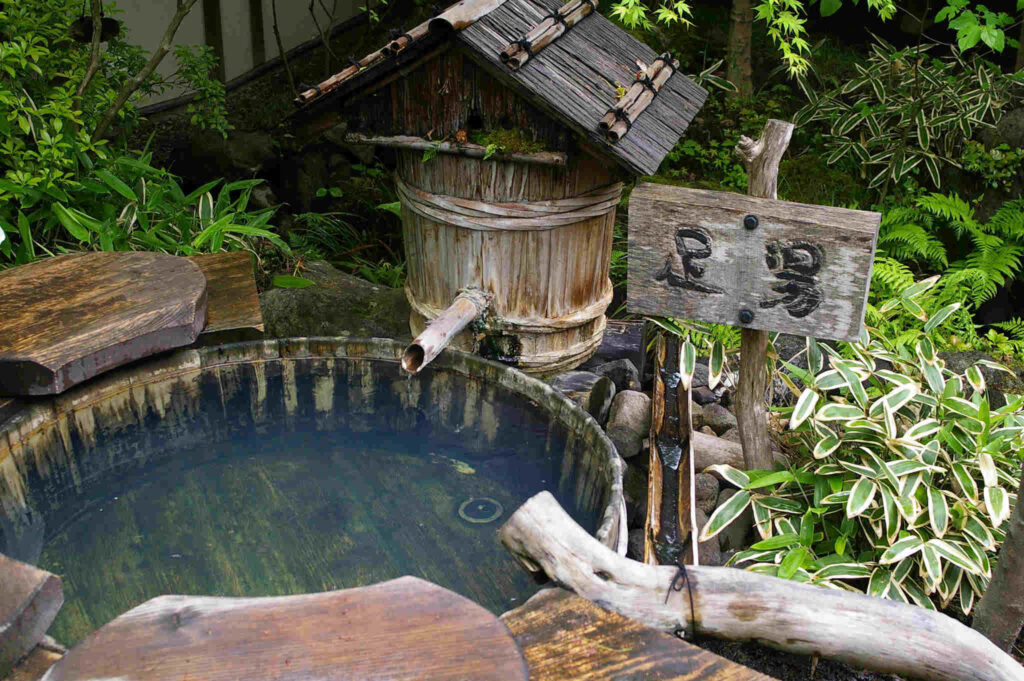 Rustikales Outdoor-Onsen im Wald mit traditionellem Holzbottich, japanischen Schriftzeichen und natürlicher Bambusvegetation für authentisches Naturerlebnis