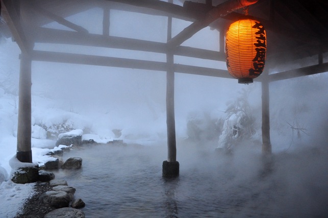 Traditionelles japanisches Onsen im Winter mit dampfendem heißem Quellwasser, rote Papierlaterne und schneebedeckte Landschaft im Hintergrund