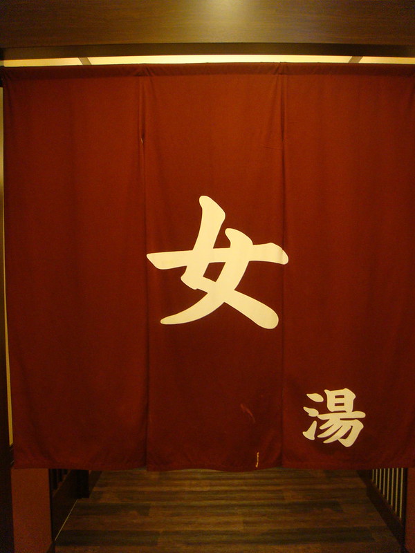 Rote Noren-Vorhänge mit japanischen Schriftzeichen "女湯" (Damenbad) am Eingang eines traditionellen Onsen-Badehauses