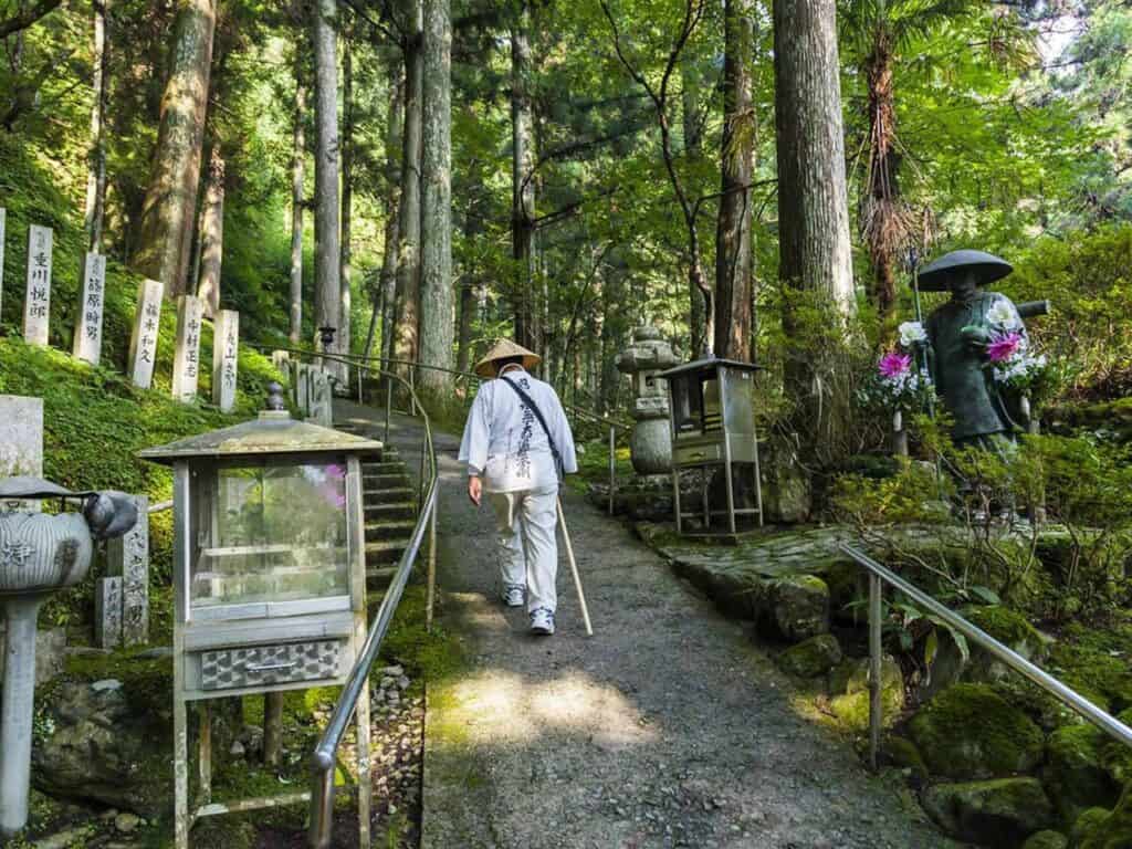 Ein Henro-Pilger in traditioneller weißer Kleidung mit Wanderstab auf einem Waldweg zwischen hohen Zedern und Steinstatuen