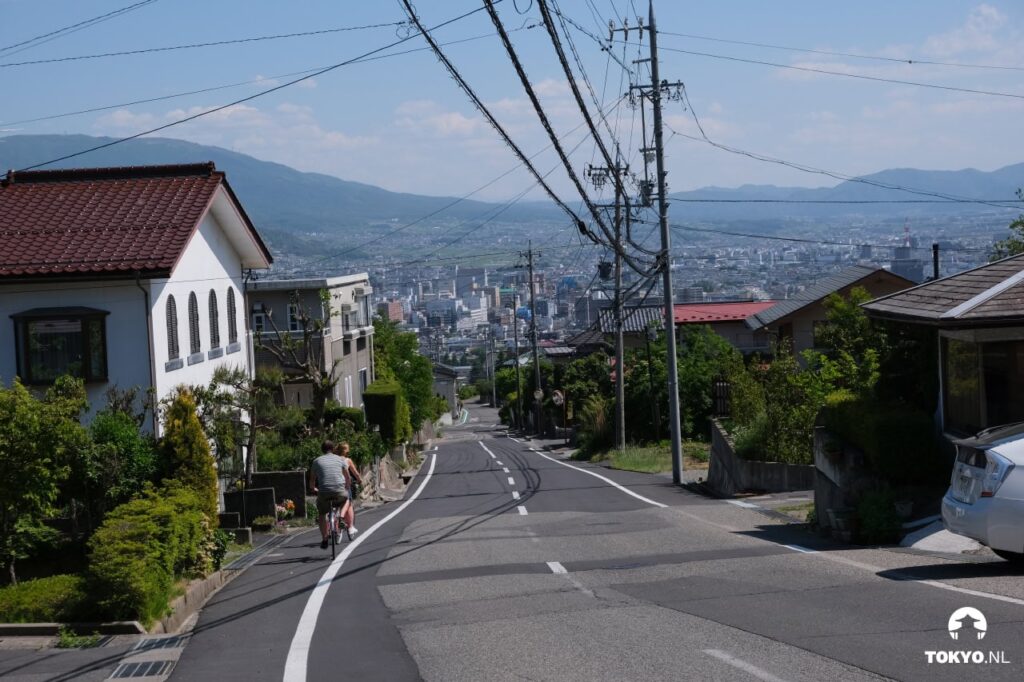 Zwei Personen auf einem Fahrrad fahren eine steile Straße in Matsumoto hinunter mit Blick auf die Stadt