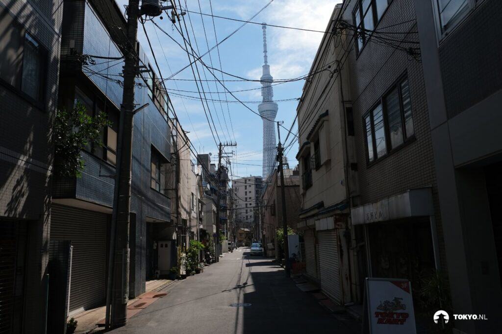Tokyo Skytree von einer schmalen Straße in Sumida aus gesehen - Der Turm erhebt sich imposant über die typischen japanischen Wohnhäuser und Stromkabel der Nachbarschaft.