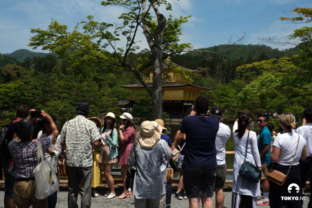 A scene showing numerous tourists gathered on a viewing platform or path, photographing and admiring the Golden Pavilion in the distance, illustrating the temple's popularity as a tourist destination.