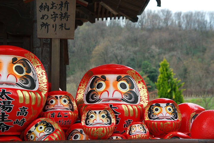 Rote Daruma-Puppen in verschiedenen Größen vor einem traditionellen japanischen Gebäude mit Bergen im Hintergrund