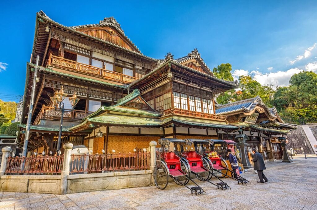 Historisches Badehaus Dōgo Onsen in Matsuyama auf der Insel Shikoku, mit traditionellen Rikschas und Holzarchitektur unter blauem Himmel