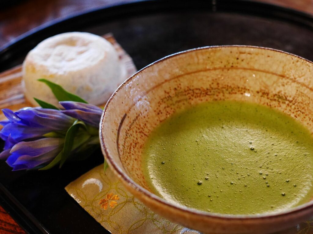 Traditionelle japanische Matcha-Teeschale mit leuchtend grünem Tee, serviert mit einer violetten Blüte und japanischen Süßigkeiten auf schwarzem Lacktablett