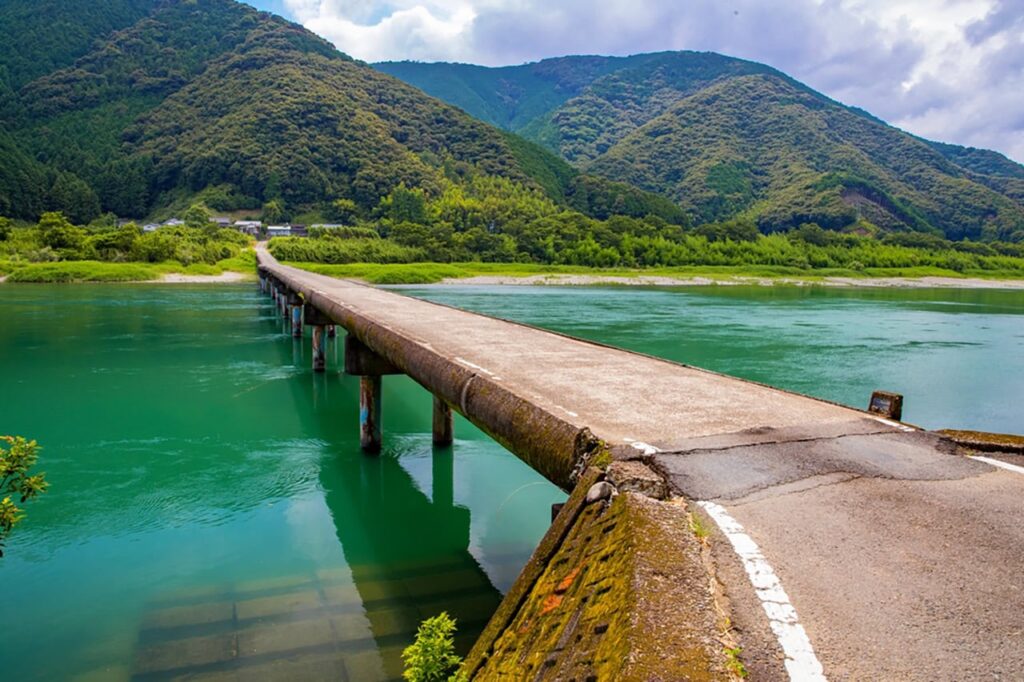 Schmale Brücke über türkisfarbenes Wasser in der Region Kōchi auf Shikoku, umrahmt von grünen Hügeln und ruhiger Natur