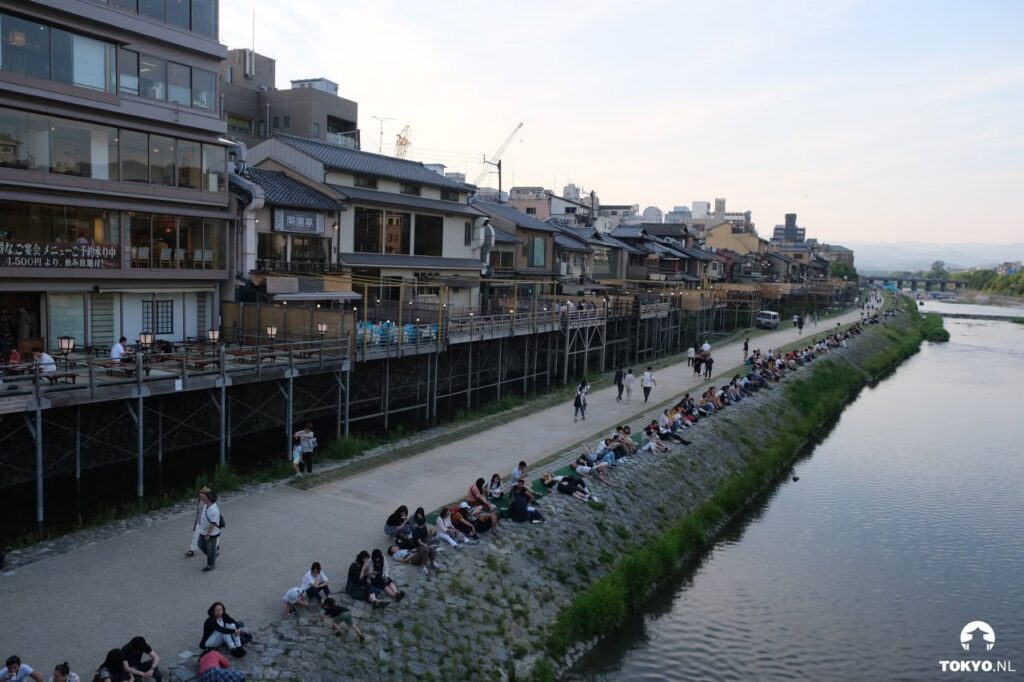 Entspannte Abendstimmung am Kamogawa-Fluss in Kyoto mit sitzenden Menschen, traditionellen Restaurants und Sonnenuntergang über der Stadt